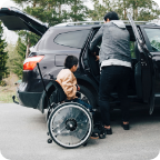 A father adjusts the car seat in his vehicle while his son waits in a wheelchair. 